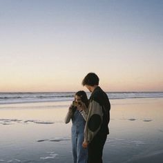 two people are standing on the beach looking at their cell phones while the sun is setting