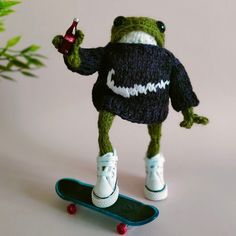 a knitted frog is standing on a skateboard and holding a drink in its hand