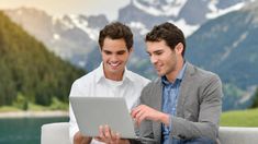 two men sitting on a couch looking at an open laptop computer in front of them