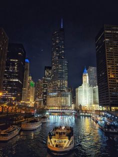 boats are floating in the water near tall buildings and skyscrapers at night with lights on