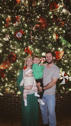 a man and woman standing in front of a christmas tree with a baby on her lap