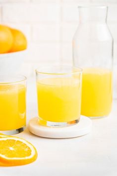 two glasses of orange juice next to an orange slice on a plate and another glass filled with orange juice