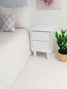 a small white nightstand next to a bed with a plant on it and a pillow