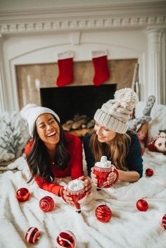 two women sitting on a bed with christmas decorations and cups in front of the fireplace