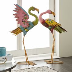 two colorful flamingos standing next to each other on a rug in front of a window