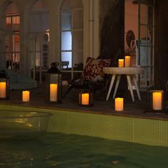 lit candles sitting on the edge of a pool in front of a table and chairs