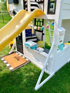 a white play house with a yellow slide in the grass next to some other toys