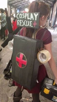 a woman is dressed up as a box with a red cross on it and holding a wrench