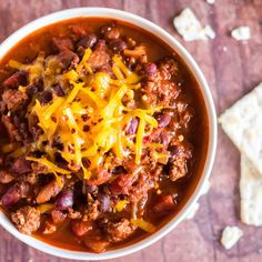 a white bowl filled with chili and cheese on top of a wooden table next to crackers