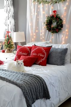 a white cat laying on top of a bed covered in red and gray pillows next to a christmas wreath