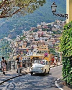 an old car is driving down the road in front of a city with lots of buildings