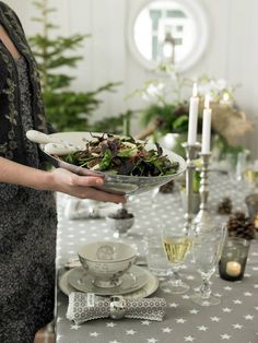 a woman holding a bowl of salad on top of a dining room table with candles in the background