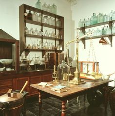 an old fashioned kitchen with lots of glass bottles on the shelves and wooden table in front of it