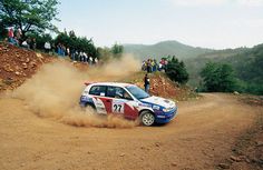 a rally car driving down a dirt road with people looking on from the sidelines