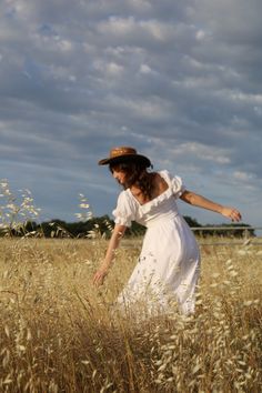 a woman in a white dress and hat walking through tall grass with her arms outstretched