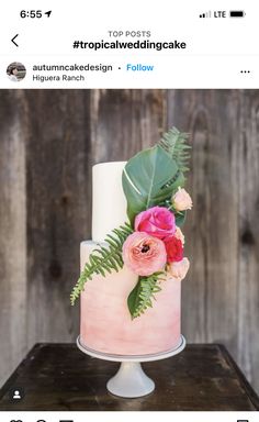 a pink cake with flowers and greenery on top sits on a table in front of a wooden wall