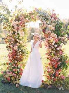 a woman in a white dress and hat standing under a floral arch with flowers on it