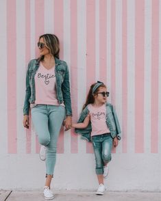 two girls wearing matching shirts and jeans leaning against a pink striped wall with their hands in each other's pockets