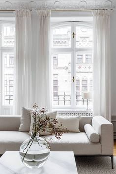 a living room filled with white furniture and large windows covered in drapes, along with a vase full of flowers on the coffee table