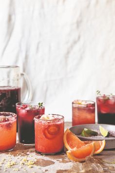 several glasses filled with different colored drinks on top of a wooden table next to sliced oranges