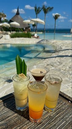 three cocktails sitting on top of a wooden table next to an outdoor swimming pool