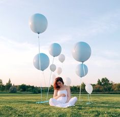 a woman sitting in the grass with balloons floating around her and an empty field behind her