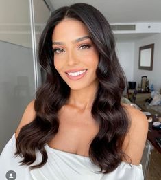 a woman with long dark hair wearing a white dress and smiling at the camera while standing in front of a mirror