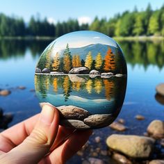 a hand holding a painted rock in front of a body of water with trees on it