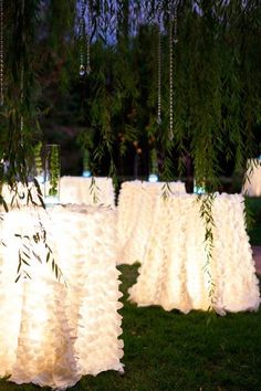 several tables covered in white ruffled cloths under a tree with lights on them