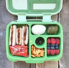 a green lunch box filled with fruit, sausages and other foods on top of a wooden table