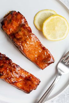 two pieces of salmon on a white plate with a fork and lemon wedges next to it