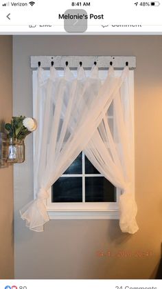 a window with white sheer curtains hanging from it's side and flowers on the ledge