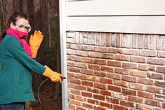 a woman in green jacket and yellow gloves holding a hose near brick wall with trees