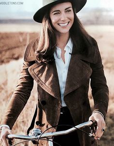 a woman wearing a hat and coat riding a bike in a field with dry grass