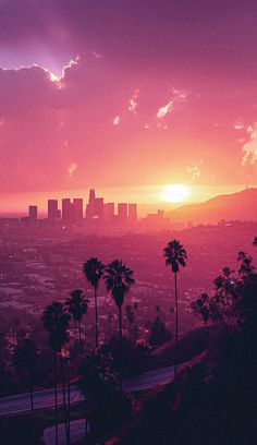 the sun is setting over a city with palm trees and mountains in the foreground