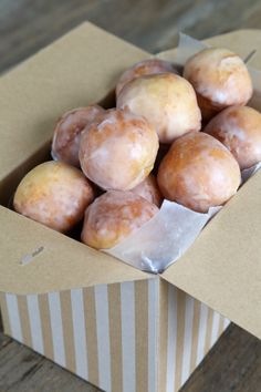 a box filled with glazed donuts sitting on top of a wooden table next to a striped paper bag