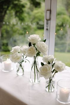 white roses in glass vases on a table with candles and greenery outside the window