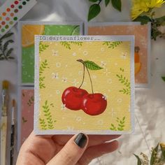 a hand holding up a card with two cherries on it and flowers in the background