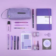 the contents of a travel bag laid out on a purple surface with pens, notebooks and other items