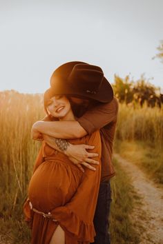 a pregnant woman hugging her husband on the side of a dirt road with tall grass in the background