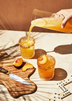 someone pouring orange juice into two glasses on top of a wooden cutting board next to sliced peaches