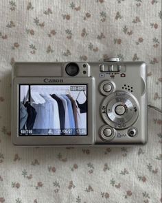 a digital camera sitting on top of a bed next to a flowered sheet with clothes hanging from it