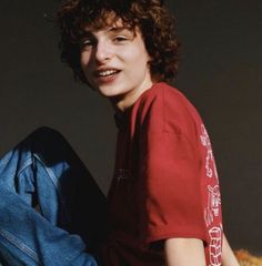 a young man with curly hair sitting on the ground wearing jeans and a red t - shirt