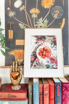 there is a painting on the wall with flowers in it and books stacked up next to each other