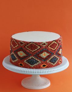 a colorful cake sitting on top of a white cake plate next to an orange wall