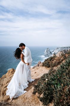 a bride and groom kissing on top of a cliff overlooking the ocean with cliffs in the background