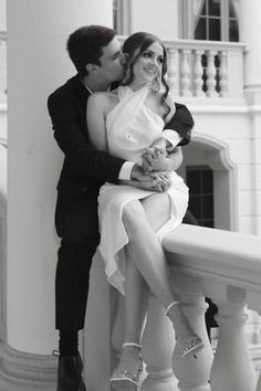 a man and woman are sitting on a balcony railing, embracing each other with their legs in the air