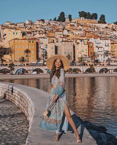 a woman in a hat and dress walking along the water