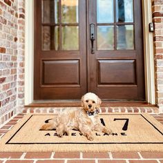 a dog sitting on the front door mat in front of a brick house with double doors