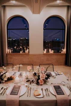 the table is set with place settings and flowers in front of two large windows at night